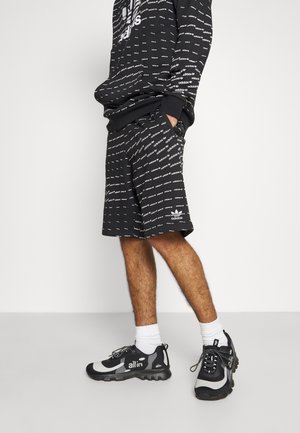 Person som bär svarta Adidas-shorts och en matchande sweatshirt med vita logotyper, vita strumpor och svarta-och-grå allin-sneakers.