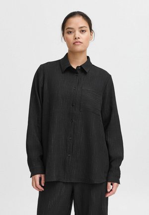 Chemise noire à manches longues avec un col, présentant un tissu texturé avec de fines rayures verticales et une poche sur le côté gauche de la poitrine.