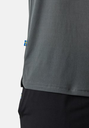 Graues, strukturiertes Sportshirt mit Karomuster und einem blauen und gelben Akzent-Tag an der Seite. Kombiniert mit schwarzen Hosen.