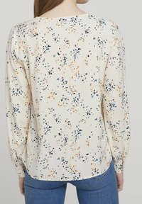 Langärmlige Bluse aus cremefarbigem Stoff mit einem mehrfarbigen, gesprenkelten Muster in Orange, Blau und Schwarz; abgerundeter Saum und leichter Glanz.