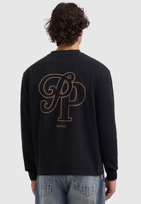 Mann mit lockigem Haar, der ein schwarzes Langarmshirt mit großen braunen "PP" und "Pure Path"-Text auf dem Rücken trägt, kombiniert mit blauen Jeans.
