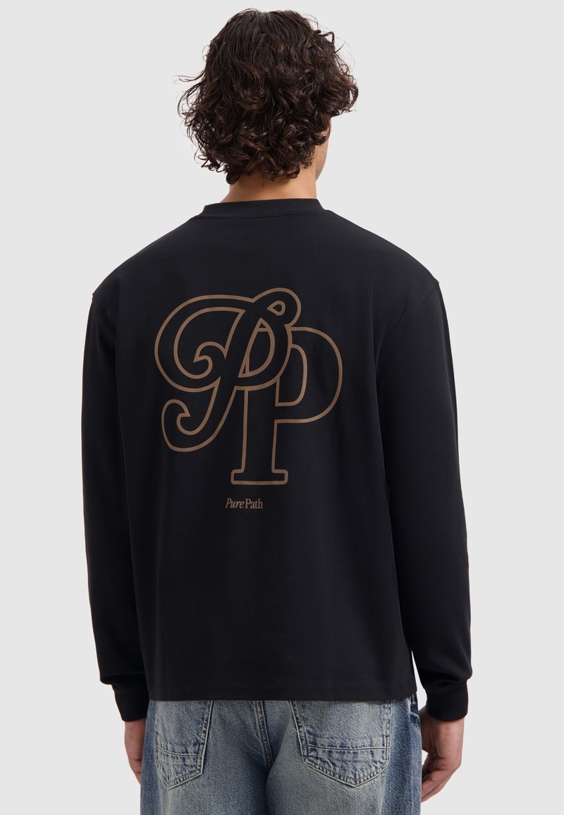 Mann mit lockigem Haar, der ein schwarzes Langarmshirt mit großen braunen "PP" und "Pure Path"-Text auf dem Rücken trägt, kombiniert mit blauen Jeans.