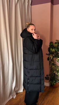 Γυναίκα με ξανθά μαλλιά μαζεμένα σε κότσο, φοράει μακρύ μαύρο μπουφάν με κουκούλα, στέκεται σε εσωτερικό χώρο κοντά σε μπεζ κουρτίνες και μια γλάστρα με φυτό.
