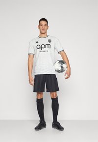 Witte voetbalshirt met zwarte afwerking en logo, gecombineerd met zwarte shorts, sokken en voetbalschoenen. Speler houdt een zwart-wit voetbalschema vast.