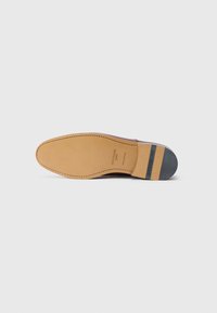 Suela de un zapato de vestir de cuero marrón con bordes cosidos y un tacón con tachuelas metálicas, marcado con la marca Heschung, sobre un fondo blanco liso.