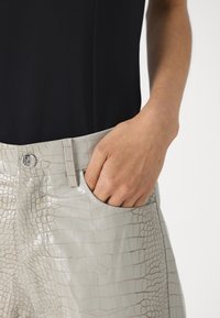 Ανοιχτό γκρι παντελόνι από τεχνητό δέρμα με ανάγλυφη υφή κροκοδείλου, με κλείσιμο κουμπιού και πλαϊνές τσέπες, σε συνδυασμό με μαύρη μπλούζα, αναδεικνύοντας λεία υφή.