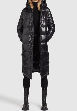 Kobieta ubrana w długą, błyszczącą czarną pikowaną kurtkę z kapturem, czarny golf, czarne legginsy i czarne botki na szarym tle.