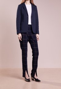 Mujer vestida con un blazzer azul marino, top blanco, pantalones de cuadros azul marino con aberturas frontales y zapatos de tacón negro, de pie sobre un suelo beige.