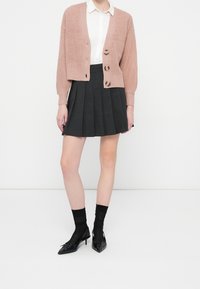 Světle růžový pletený kardigan se třemi knoflíky, doplněný skládanou tmavě šedou sukní. Černé lodičky na vysokém podpatku a černé ponožky dokončují outfit.