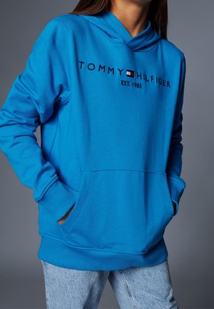 Kobieta w jasnoniebieskiej bluzie z kapturem Tommy Hilfiger z rękami w przedniej kieszeni, ubrana w jasnoniebieskie dżinsy na szarym tle.