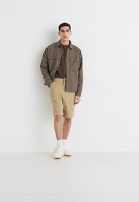 Brown button-up jacket over a brown t-shirt, paired with tan shorts and white sneakers, against a plain white background.
