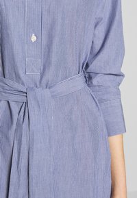 Modrá a bílá pruhovaná košile s límečkem na knoflíky, opaskem v pase a dlouhými rukávy s knoflíkovými manžetami a přizpůsobeným střihem.