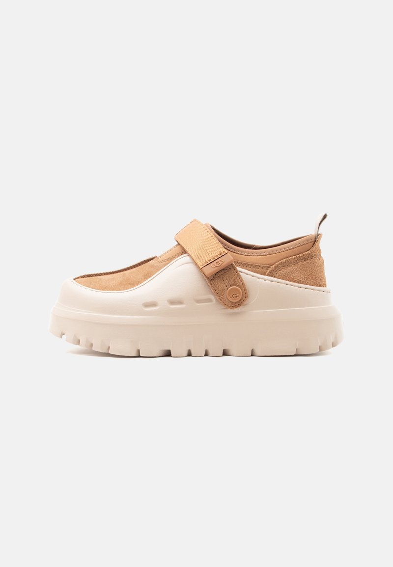 Chaussure beige et marron clair à enfiler, avec semelle épaisse à crampons et bride réglable sur le dessus, composée de matériaux en suède et en caoutchouc.