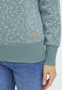 Felpa verde con puntini bianchi, blu e beige sparsi; manicotti e orlo a coste; presenta un piccolo tag logo beige sul davanti.