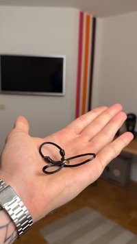 Fekete zsinór karkötő, amely hurkos masni mintával rendelkezik, és egy kis arany díszítés rögzíti. Egy kéz tartja a karkötőt, kiemelve annak kompakt méretét.