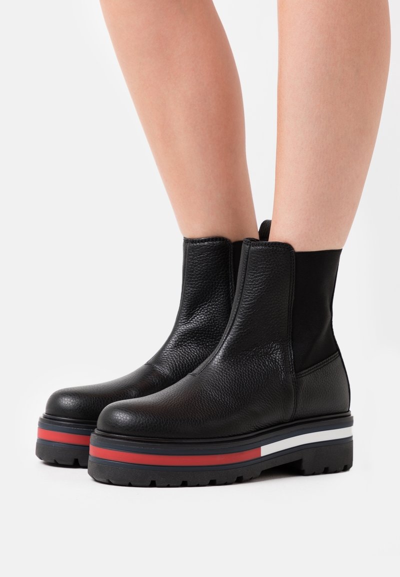 Bottines en cuir noir avec des semelles épaisses rayées rouge, blanc et noir, portées sur des jambes nues, présentées sur un fond blanc.
