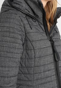 Grijze gewatteerde jas met subtiel ruitpatroon, zwarte rits en capuchon met trekkoorden, gedragen door een persoon met lichtbruin haar.