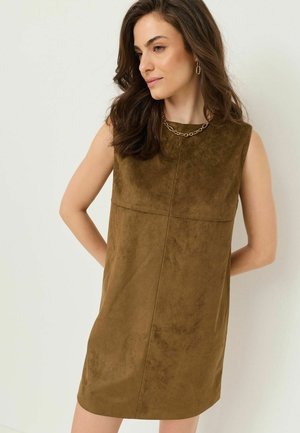 Frau mit welligem braunem Haar, die ein ärmelloses olivgrünes Wildlederkleid und eine goldene Kettenhalskette trägt und mit den Händen hinter dem Rücken posiert.