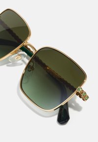 Kvadrātveida saulesbrilles ar zaļiem gradienta stikliem, zelta metāla rāmī un dekoratīviem akcentiem, kuros ir trīs zaļas akmeņi uz tempļiem.