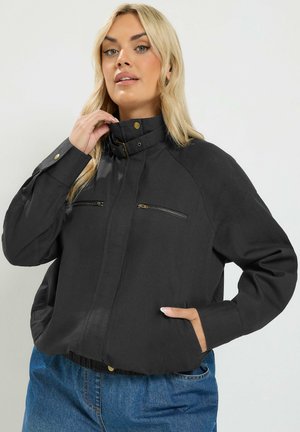 Lyshåret kvinde iført sort lynlåsflyverjakke med høj krave og blå jeans, den ene hånd justerer kraven, den anden i lommen, neutral baggrund.