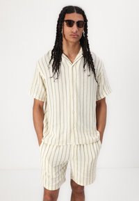 Camicia a maniche corte a righe color crema e pantaloni abbinati, realizzati in un tessuto leggero e strutturato. Include bottoni e colletto.