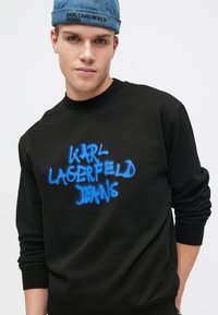 Svart hettegenser med blå graffiti-stil "KARL LAGERFELD JEANS" logo. Rund hals, lange ermer og ribbede mansjetter. Solid, myk tekstur.