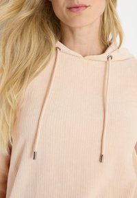 Femme blonde portant un sweat à capuche côtelé rose clair avec des cordons, visible des épaules aux lèvres sur fond blanc.