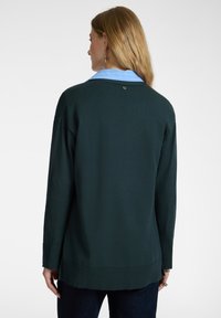 Nainen, jolla on pitkät vaaleat hiukset, pukeutuneena tumman vihreään neulepaitaan vaaleansinisen kauluspuseron yli, nähtynä takaapäin yksinkertaista valkoista taustaa vasten.