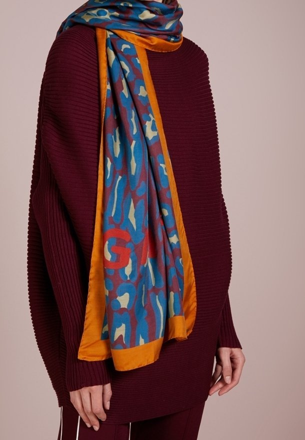 Personne portant un pull côtelé rouge foncé avec une écharpe colorée bleu, orange et rouge à motifs drapée sur l'épaule sur un fond uni.
