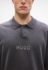 Sötét szürke póló, texturált bordás gallérral, egygombos záródással és hímzett "HUGO" logóval a mellrészen.