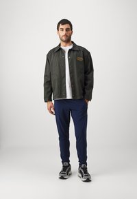 Donkergroene corduroy jas met geel logo, witte t-shirt eronder, marineblauwe joggers en zwart-grijze sportschoenen.