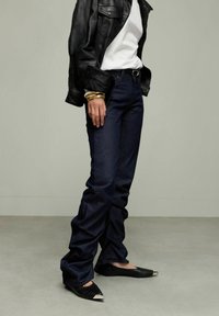 Jeans droit en denim foncé, associés à un t-shirt blanc et une veste en cuir noire. Les chaussures noires présentent des bouts métalliques pointus. Des bracelets en or mettent en valeur le poignet.