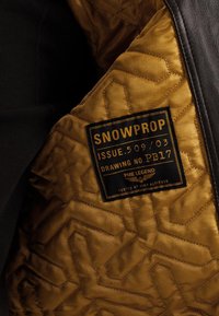 Brun quiltet interiør af en jakke med en sort læderoverflade. Har et mærke, der læser "SNOWPROP," med metalaccenter og et geometrisk mønster.