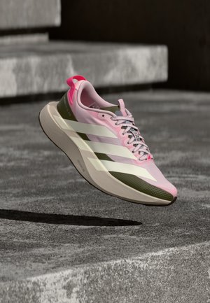 Pink-grüner Laufschuh mit weißen Streifen und strukturierter Sohle, der über Betonstufen im Außenbereich schwebt.