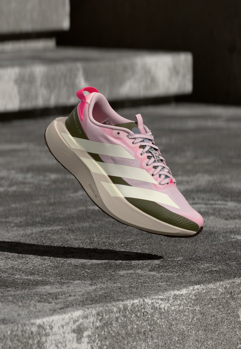 Chaussure de course rose et verte avec des bandes blanches et une semelle texturée flottant au-dessus de marches en béton dans un cadre extérieur.