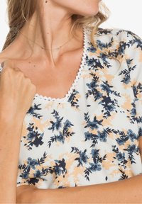Blusa floral en blanco con patrones en azul marino y melocotón, con un escote scalloped y mangas cortas. Textura de tela suave.