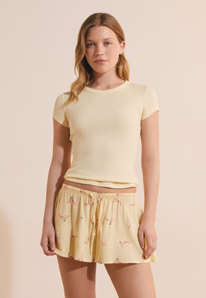 Joven mujer que lleva una camiseta de manga corta acanalada de color amarillo pálido y pantalones cortos amarillos a juego con estampado de pájaros rosas, de pie frente a un fondo liso.
