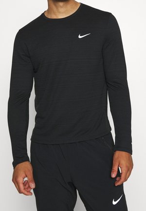 Mann trägt ein schwarzes langärmeliges Nike-Shirt und schwarze Nike-Hose mit sichtbaren weißen Swoosh-Logos auf Brust und Oberschenkel.