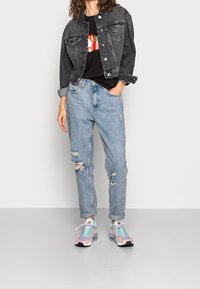 Svart denimjacka, grafisk svart t-shirt, slitna ljusblå jeans och flerfärgade sneakers med pastellaccent och svarta sulor.
