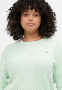 Mintgrønn kabelstrikket genser med rund hals, med teksturerte mønstre og et lite logoakcent på brystet. Mykt stoff.