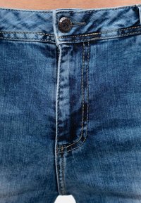 Jean en denim à taille mi-haute, couleur bleue, avec des coutures visibles et un bouton en métal, présentant une texture délavée et une coupe classique.