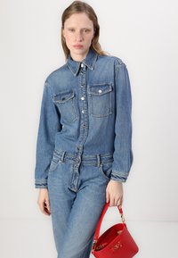 Τζιν ολόσωμη φόρμα σε ανοιχτό μπλε χρώμα με γιακά, κουμπιά μπροστά και δύο τσέπες στο στήθος; συνδυασμένη με μια μικρή κόκκινη τσάντα.