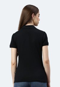 Frau mit langen braunen Haaren, die ein schwarzes, kurzärmeliges Poloshirt und blaue Jeans trägt, mit dem Rücken zum Betrachter vor einem einfarbigen hellen Hintergrund.