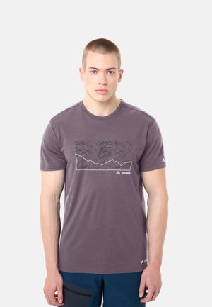 Giovane uomo con capelli corti che indossa una maglietta viola con una grafica a linea contorno di montagna e pantaloncini blu, in piedi contro uno sfondo semplice.