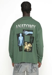 Grøn langærmet t-shirt med et grafisk design, der viser anatomiske illustrationer og blomsterakcenter på ryggen. Sølvfarvet tekst står der "ANATOMY."