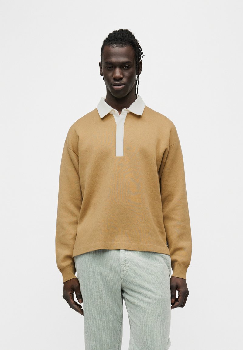 Χακί βαμβακερό πουλόβερ με λευκό γιακά, διαθέτει σχέδιο με ημιδιάφανο κουμπί και ριμπ μανσέτες, συνδυάζεται με ανοιχτό πράσινο παντελόνι με υφή.