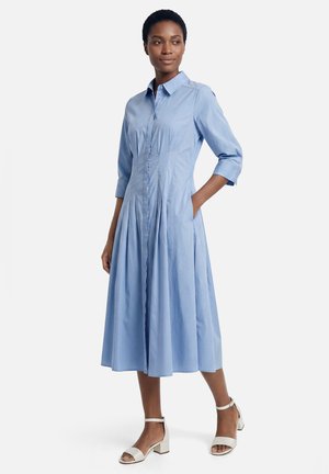 Frau trägt ein hellblaues, knöpfbares Midikleid mit Dreiviertelärmeln und weißen Sandalen mit Blockabsatz und Knöchelriemen.