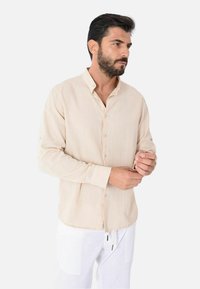 Camicia a maniche lunghe beige chiaro con colletto a punta, bottoni frontali e una texture morbida. Indossata con pantaloni bianchi dotati di coulisse.