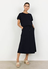 Dunkelblaues Kleid mit kurzen Ärmeln, gebundenem Taillenbund und zwei seitlichen Taschen, kombiniert mit beigen, gewebten Sandalen mit Blockabsatz. Glattes, lässiges Material.