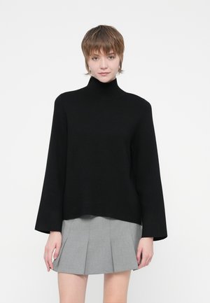 Črna turtleneck pulover s dolgimi rokavi in sproščenim krojem, izdelan iz mehkega materiala, v kombinaciji s sivo naborkano krilo.